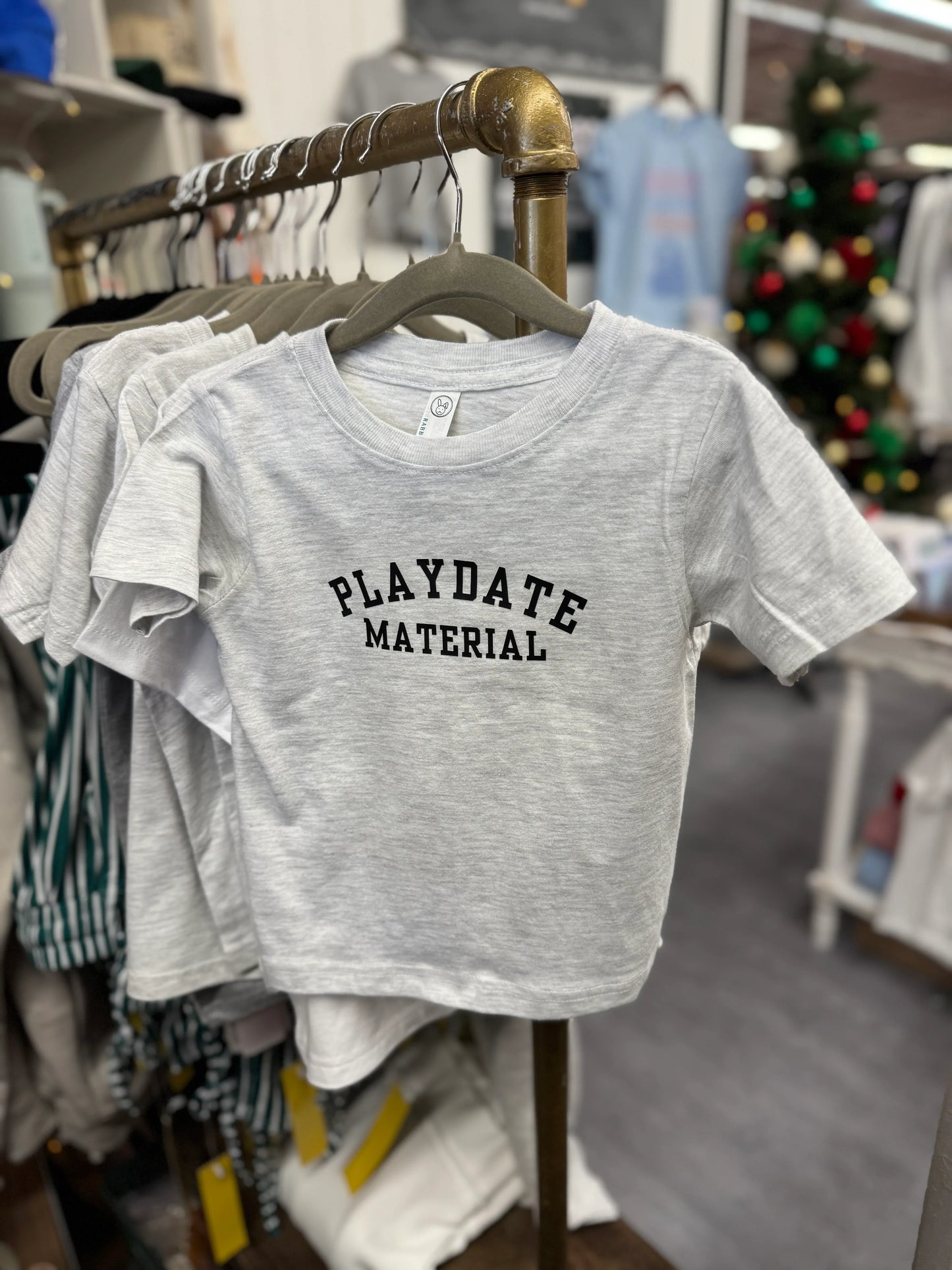 Gray t-shirt with 'Playdate Material' text on a clothing rack in a store.
