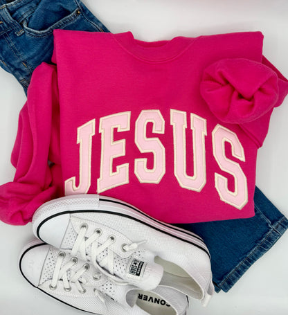Pink sweatshirt with 'JESUS' text, white sneakers, and blue jeans on a white background