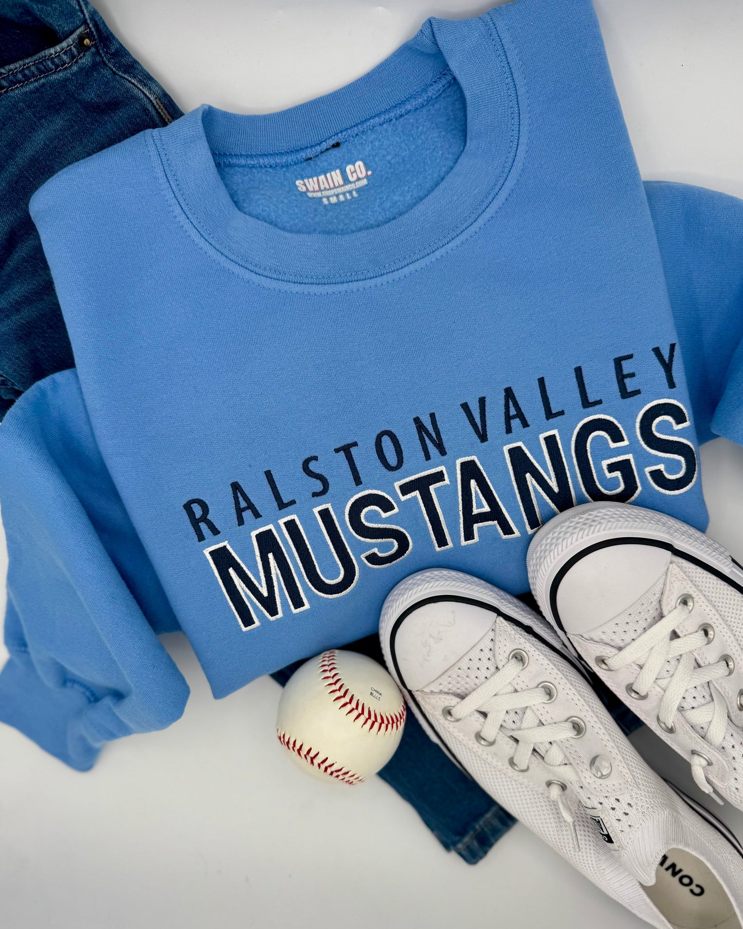 Blue sweatshirt with 'Ralston Valley Mustangs' text, white sneakers, and baseball on a white surface.