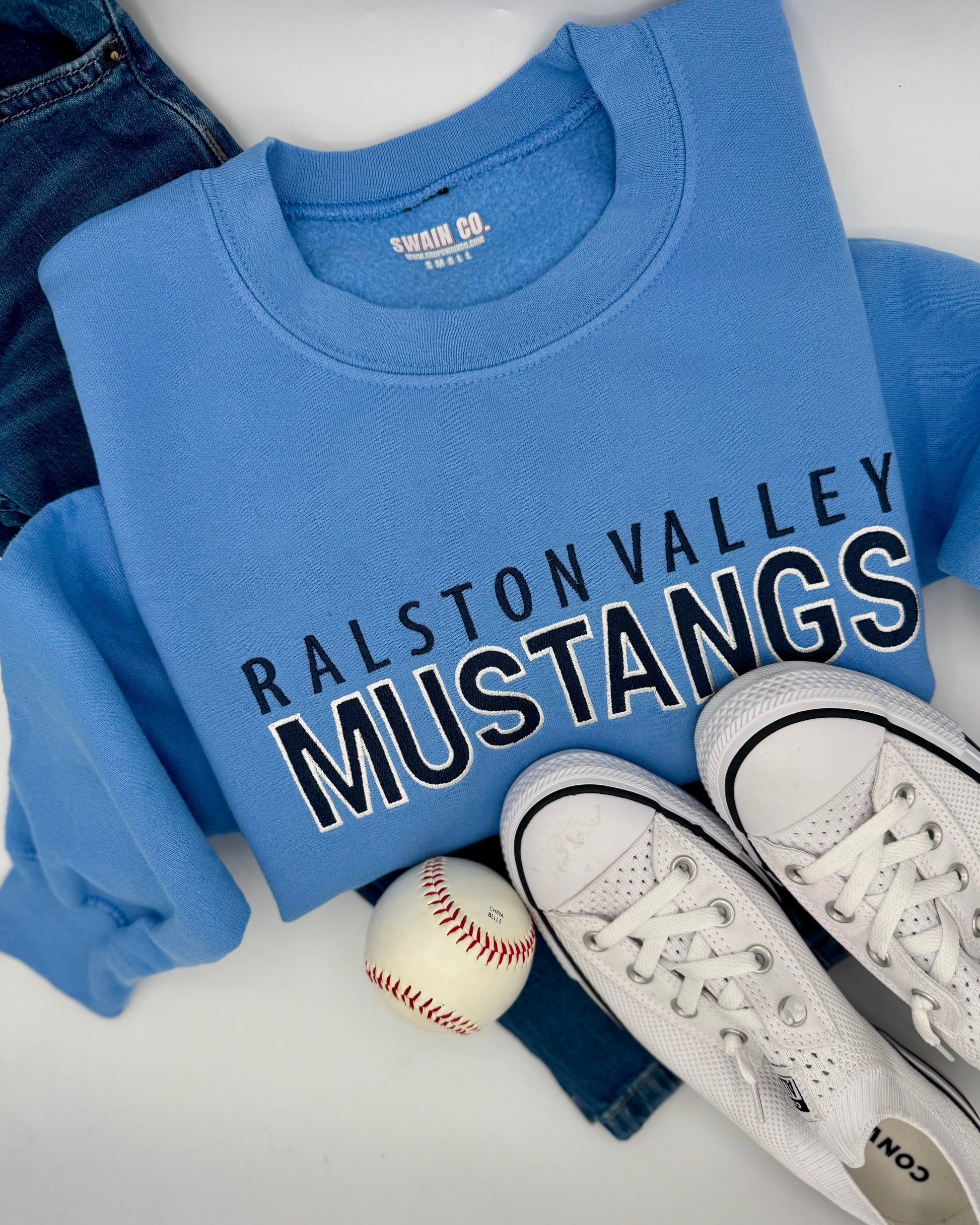 Blue sweatshirt with 'Ralston Valley Mustangs' text, white sneakers, and baseball on a white surface.