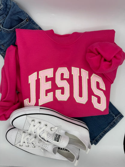 Pink sweatshirt with 'JESUS' text, white sneakers, and denim jeans on a light background