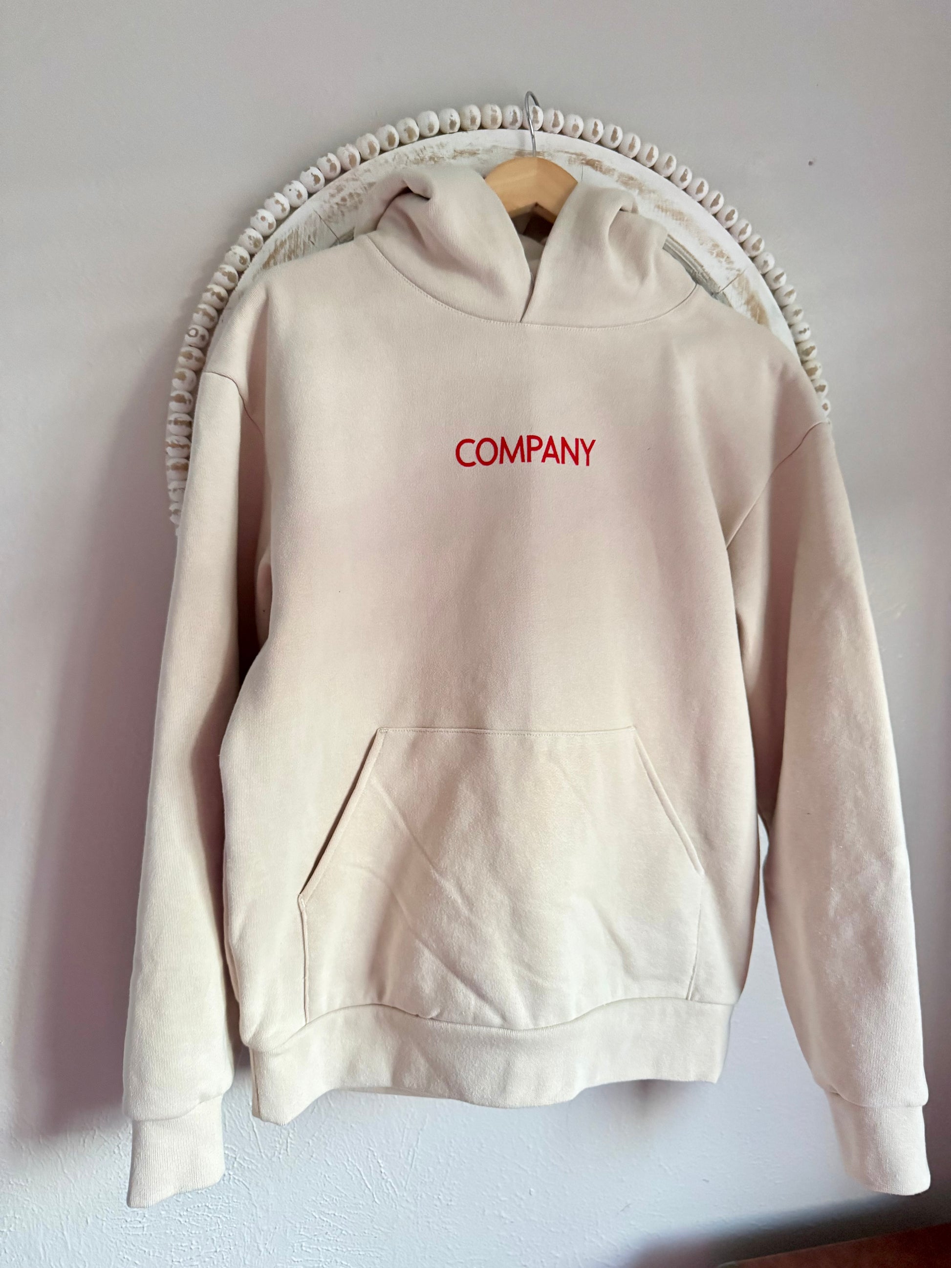 Beige hoodie with 'COMPANY' text in red on a wooden hanger against a light gray wall.