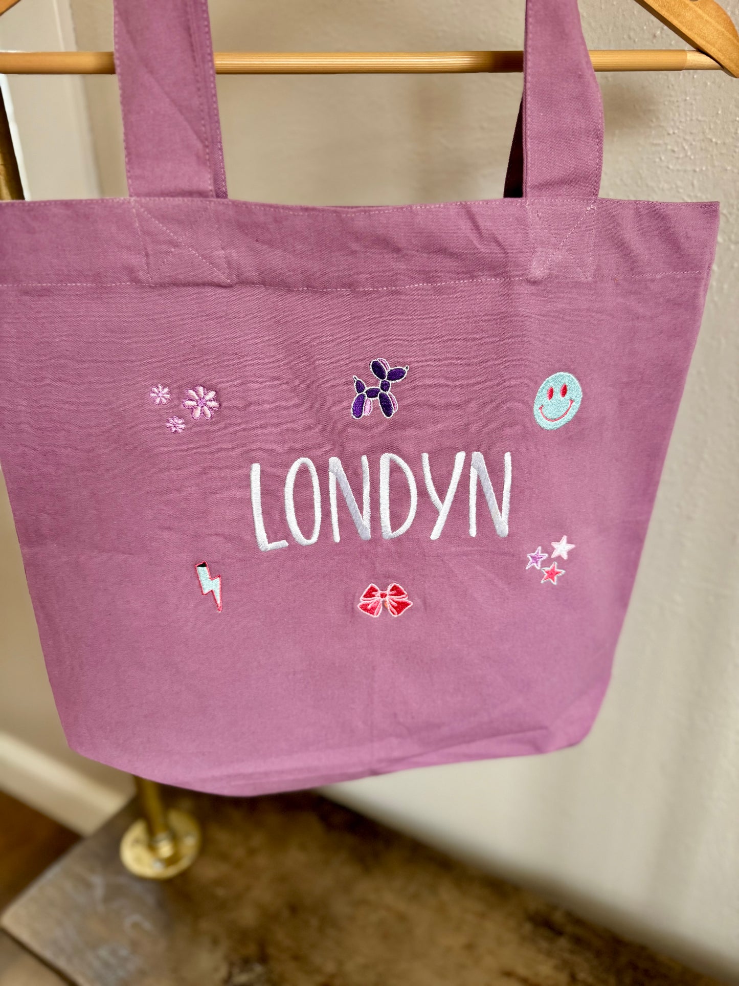 Purple tote bag with 'LONDYN' and decorative elements on a wooden rack.