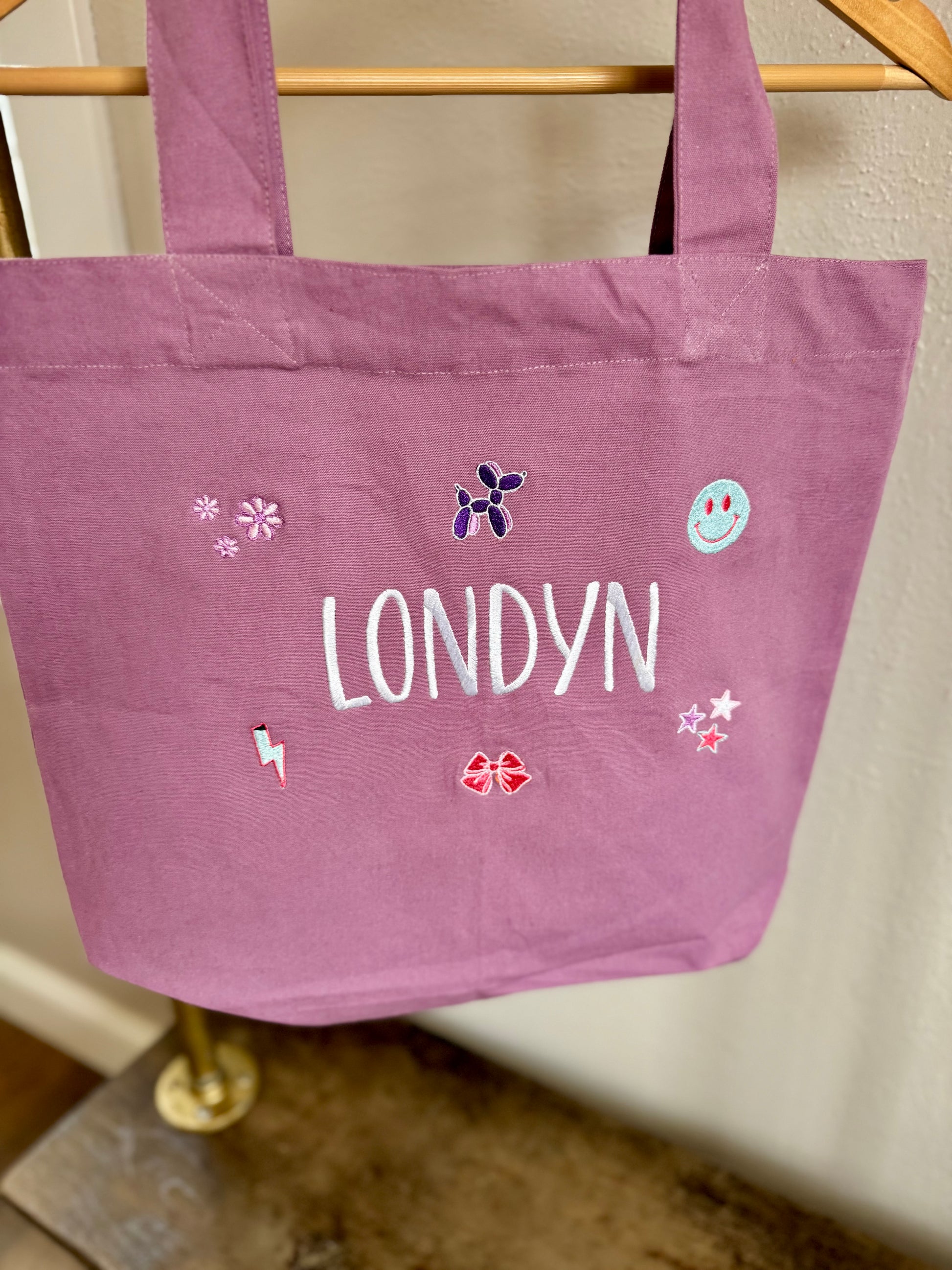 Purple tote bag with 'LONDYN' and decorative elements on a wooden rack.