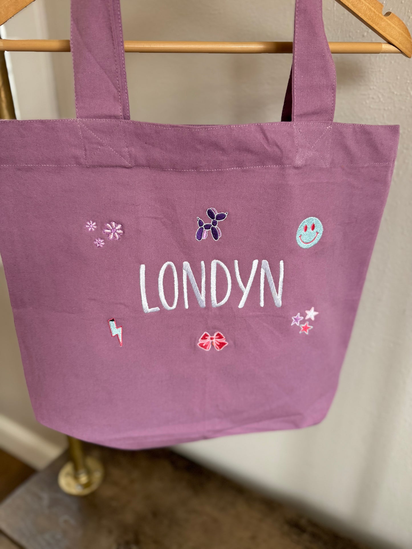 Purple tote bag with 'LONDYN' and decorative elements on a wooden rack.