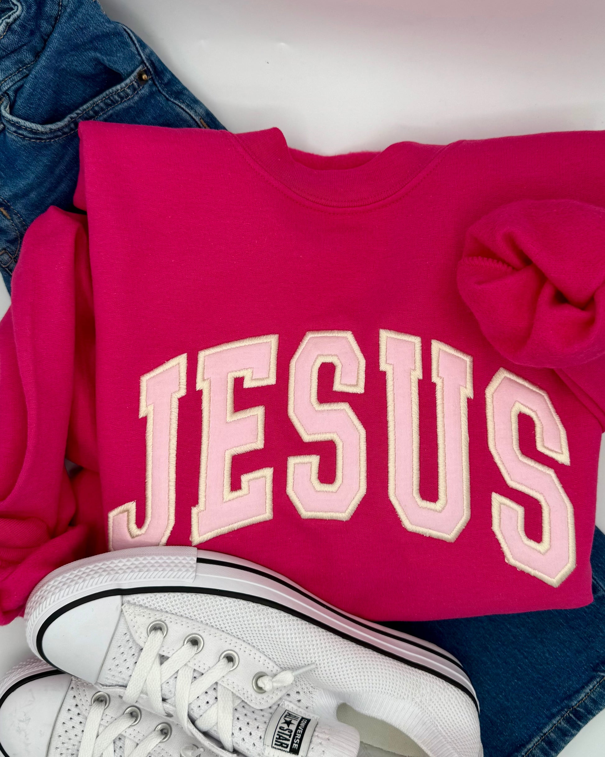 Pink sweatshirt with 'JESUS' printed on it, paired with white sneakers and blue jeans.