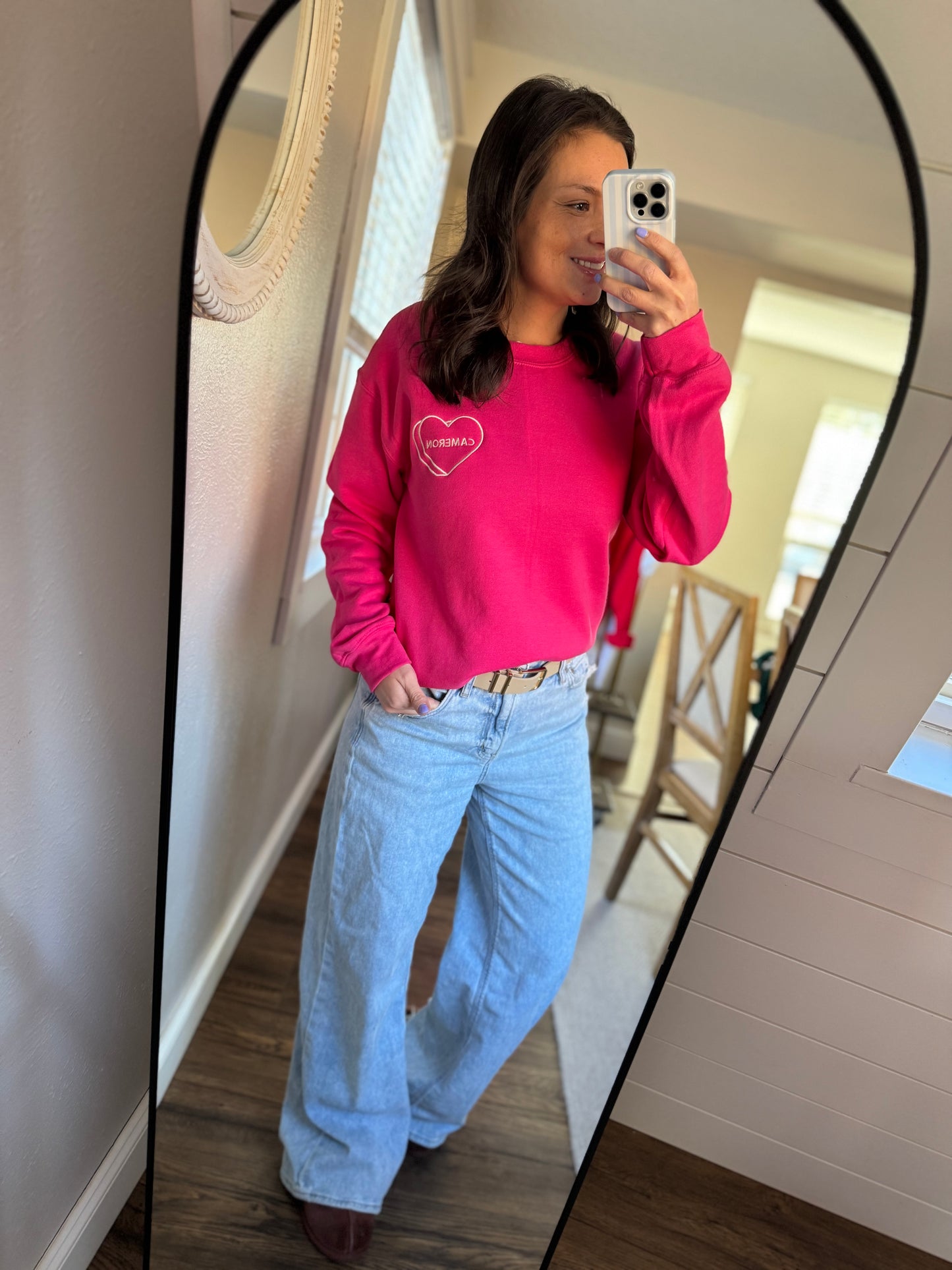 Person wearing a pink sweatshirt with a heart design and light blue jeans, standing in front of a mirror.