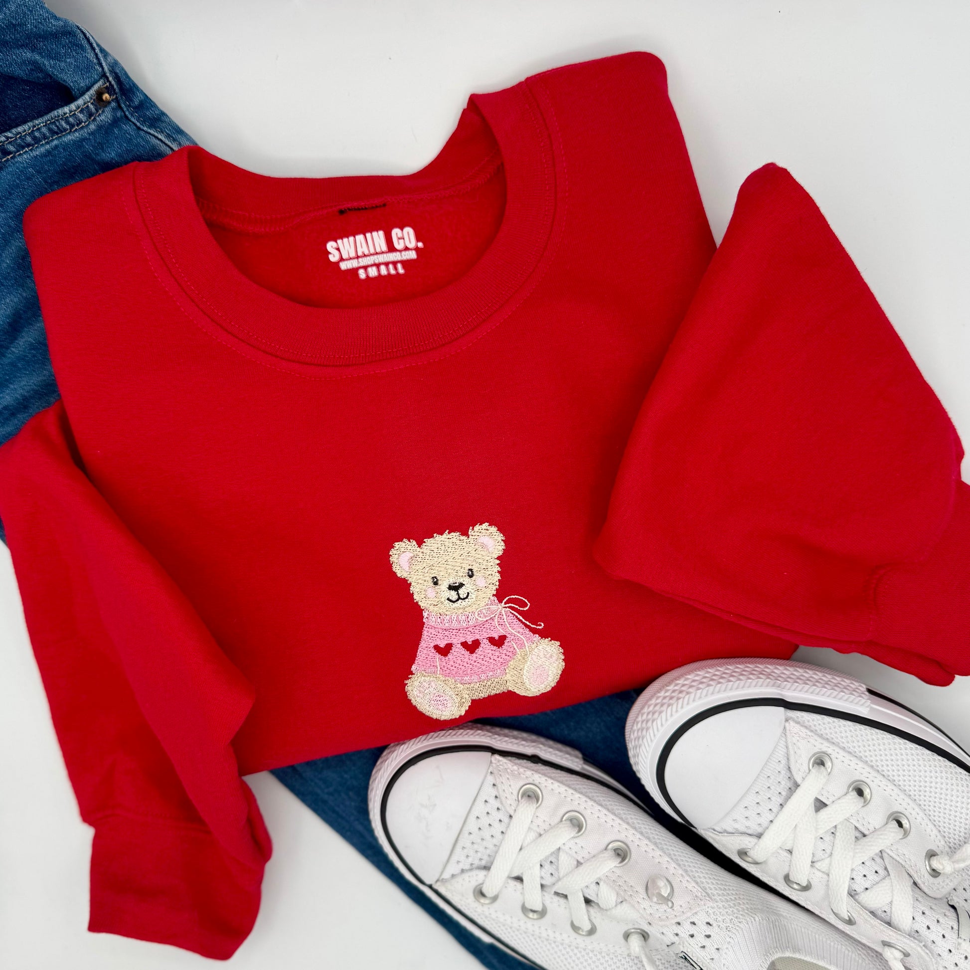 Red sweatshirt with teddy bear design, white sneakers, and blue jeans on a white background
