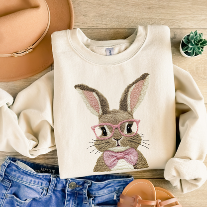 Beige sweatshirt with a faux embroidered bunny design wearing glasses and a bow tie, placed on a wooden surface with jeans and sandals.