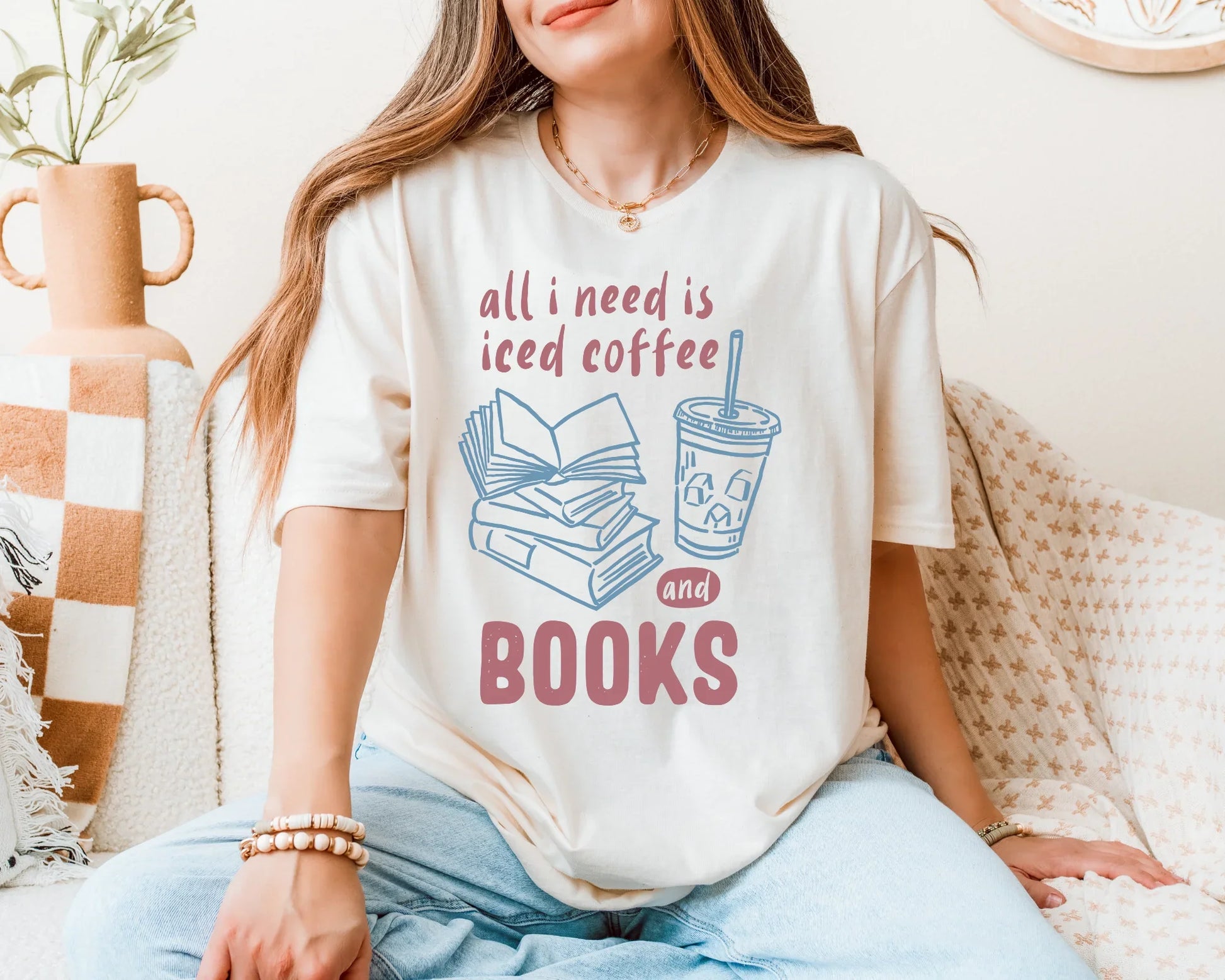 Person wearing a t-shirt with 'all I need is iced coffee and books' design, sitting on a couch.