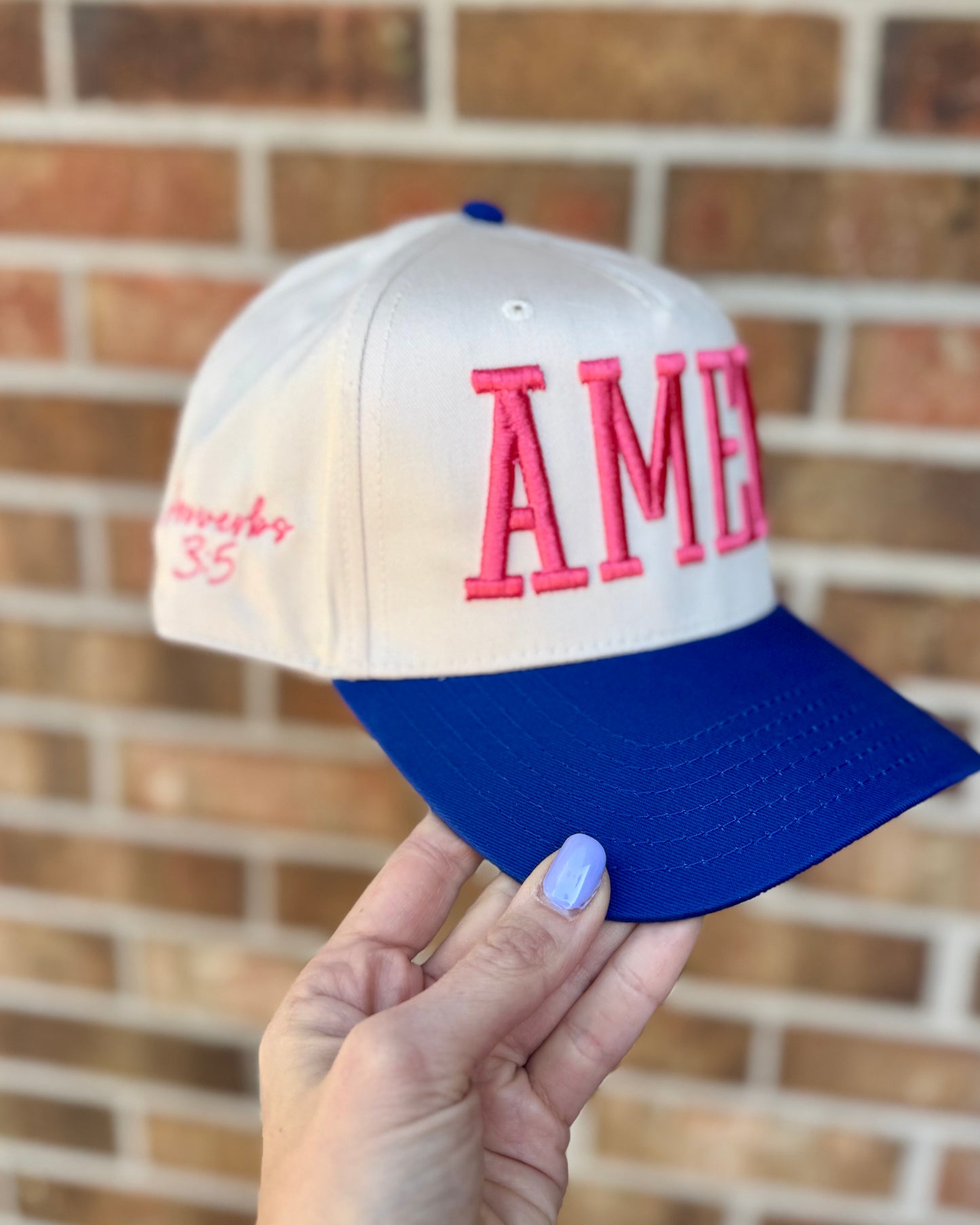 Cap with 'AMEN' embroidered in pink letters held by a hand against a brick wall.