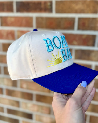 White and blue cap with 'BOAT BAY' text held against a brick wall.