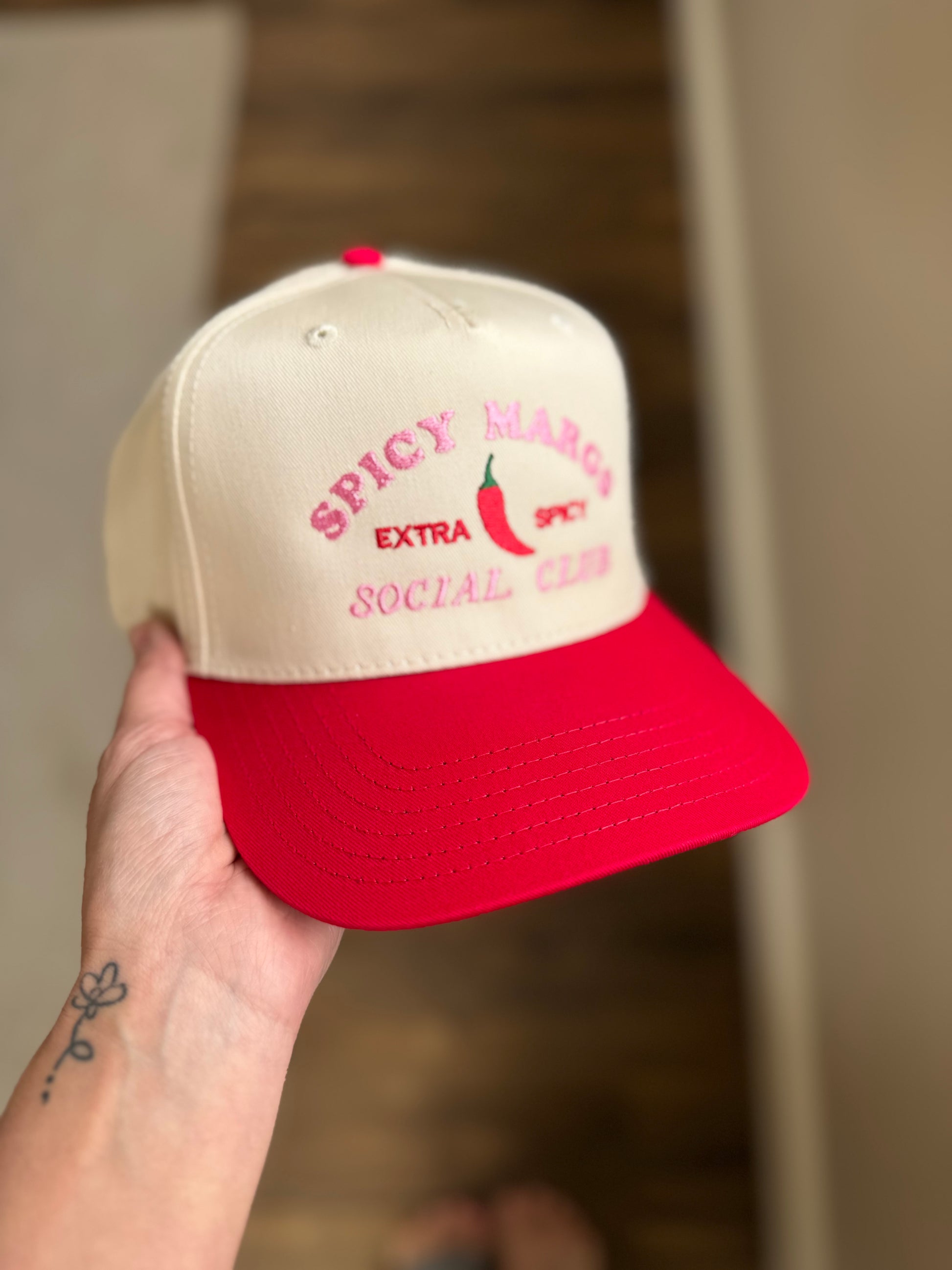 Beige and red cap with 'Spicy Mango Extra Social Club' text held by a hand.