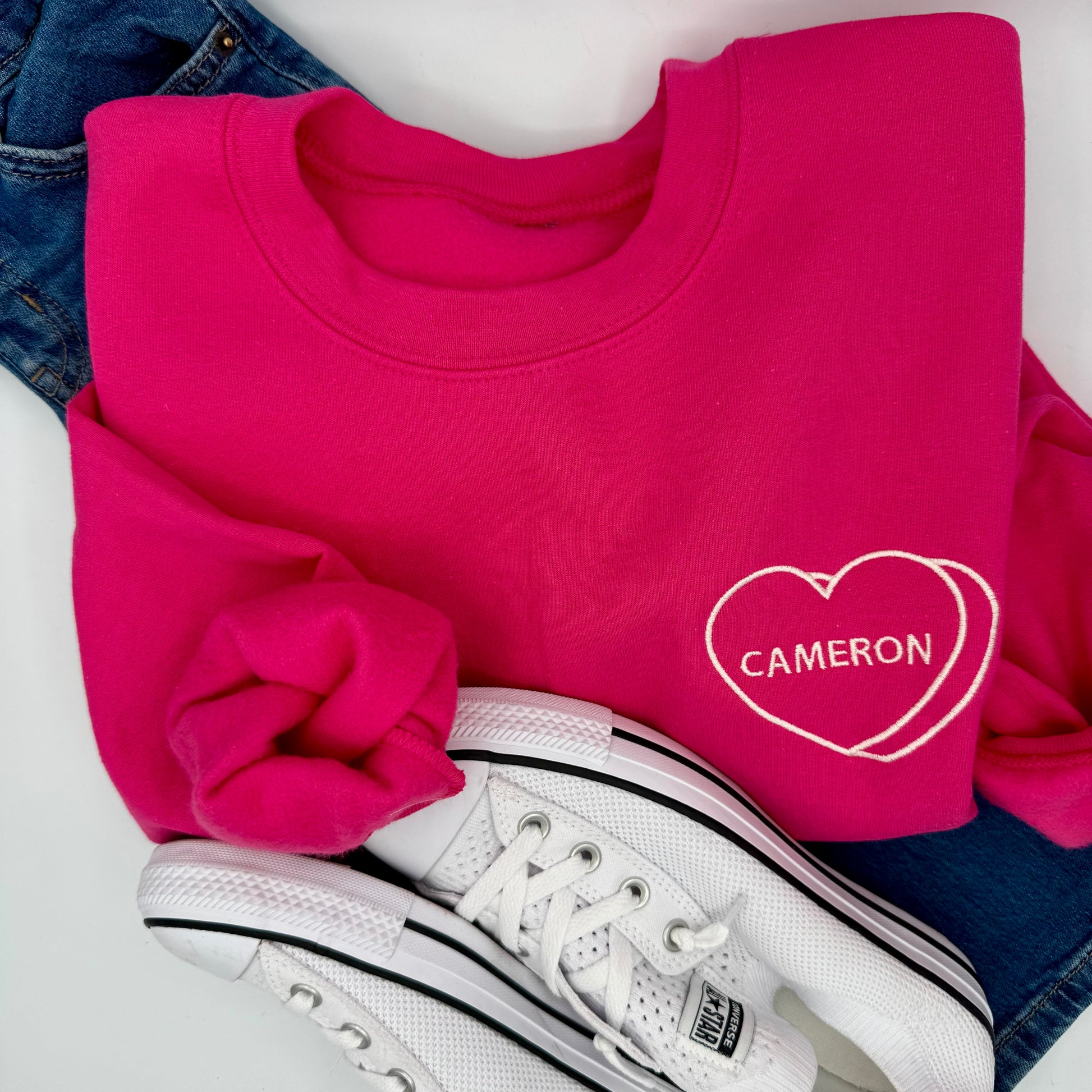 Pink sweatshirt with 'Cameron' design, white sneakers, and blue jeans on a white surface