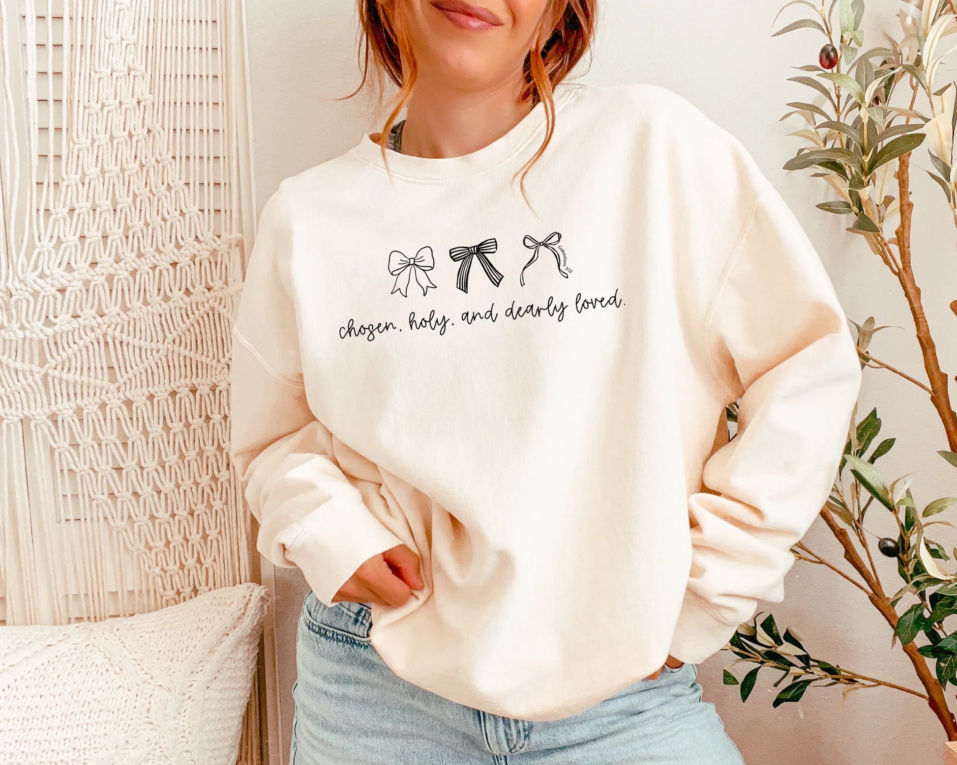 Person wearing a cream sweatshirt with text and bow graphics, standing indoors.