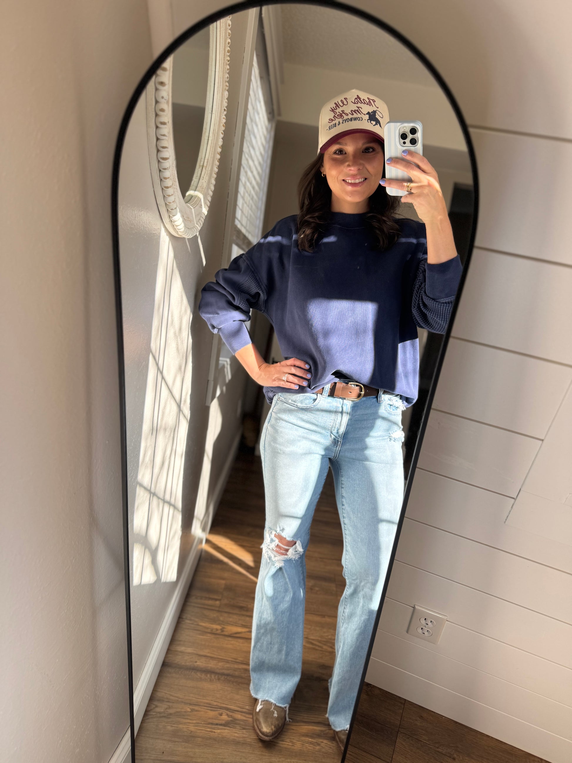 Person taking a mirror selfie wearing a blue sweater, light blue jeans, and a beige cap.