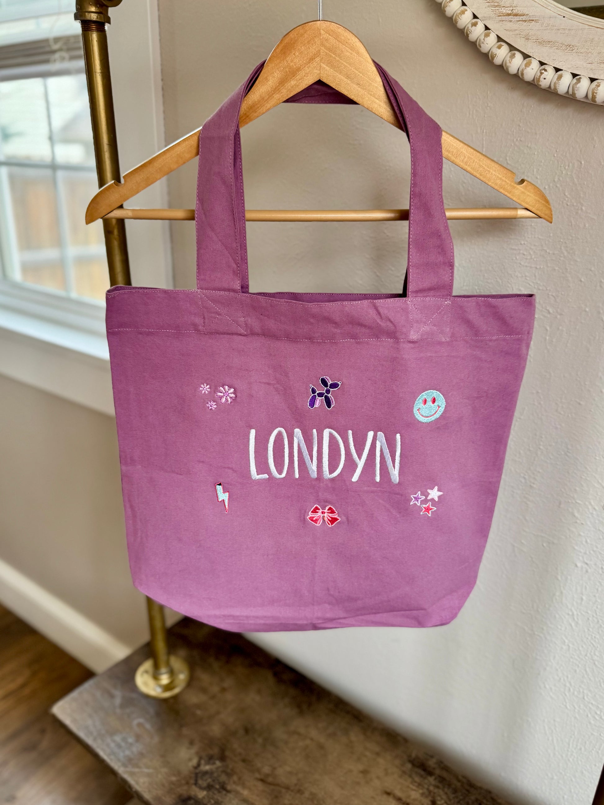 Purple tote bag with 'LONDYN' printed on it, hanging on a gold hanger against a neutral wall.