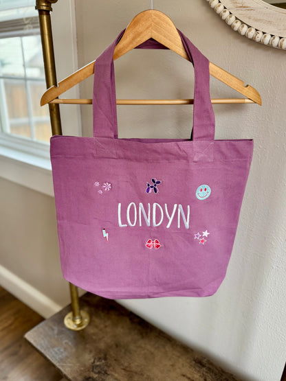 Purple tote bag with 'LONDYN' printed on it, hanging on a gold hanger against a neutral wall.
