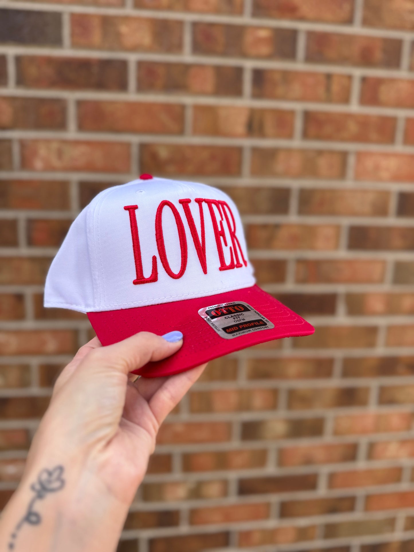 White and red cap with 'LOVER' text held against a brick wall