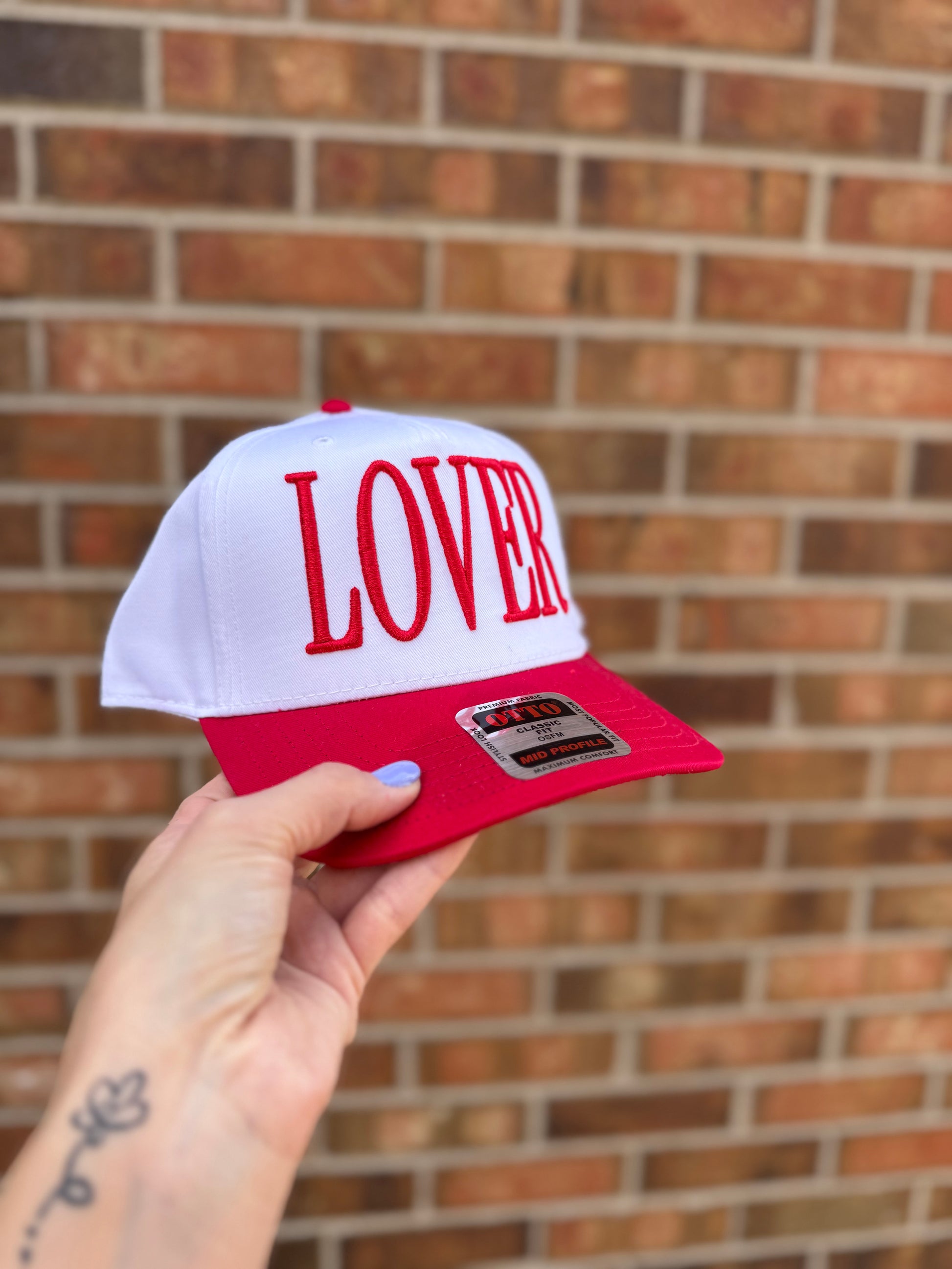 White and red cap with 'LOVER' text held against a brick wall