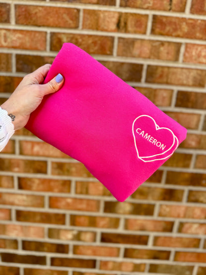 Pink sweatshirt with 'Cameron' embroidered with a heart, held against a brick wall.