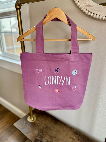 Purple tote bag with 'LONDYN' printed on it, hanging on a wooden hanger against a neutral wall.