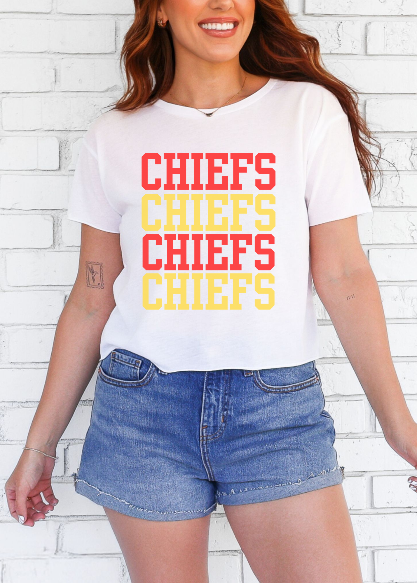 Person wearing a white t-shirt with 'CHIEFS' printed in red and yellow, standing against a white brick wall.