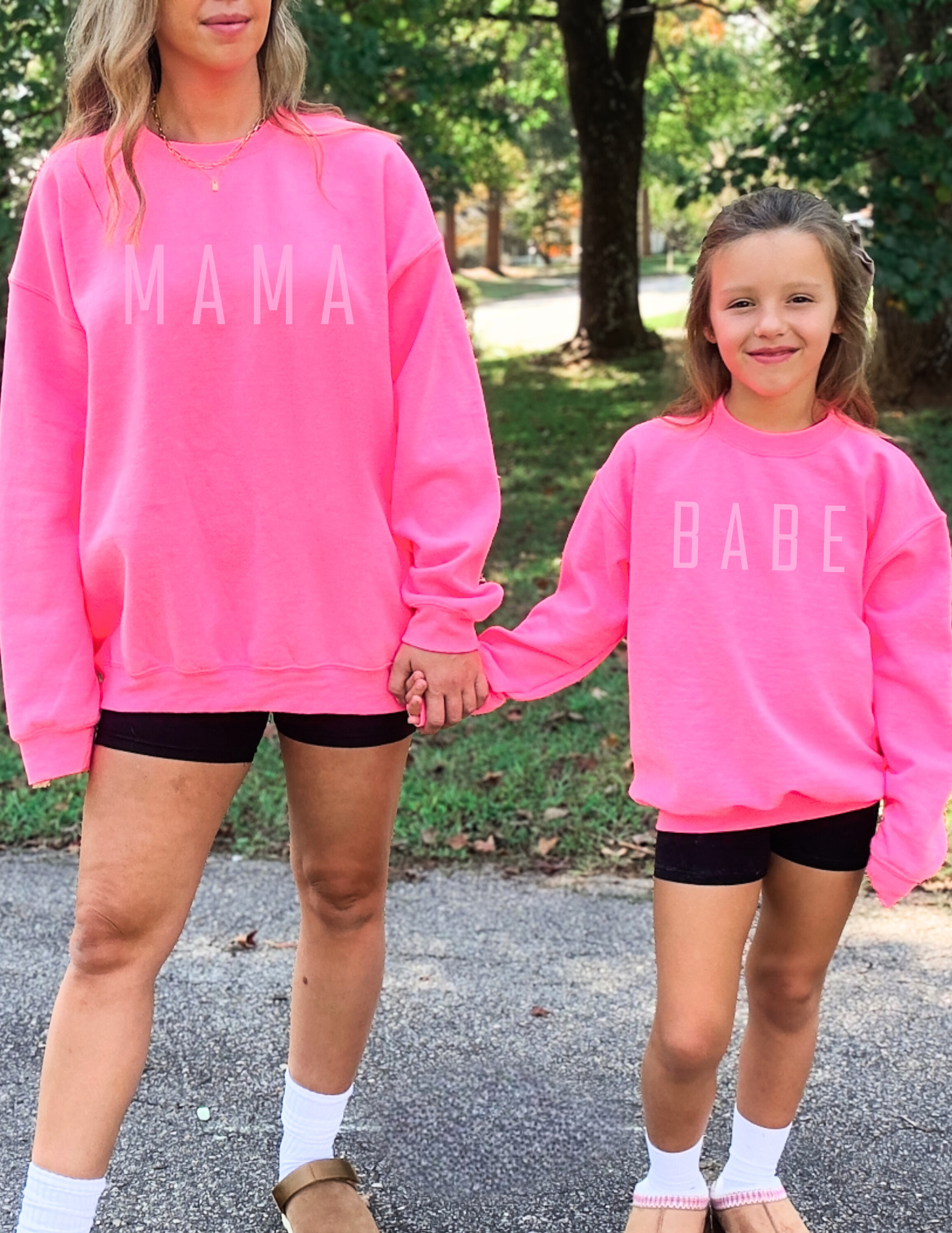 Two people wearing pink sweatshirts with 'MAMA' and 'BABE' text, standing outdoors.