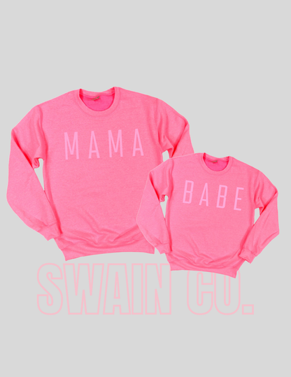 Two pink sweatshirts with 'MAMA' and 'BABE' text on a light gray background.