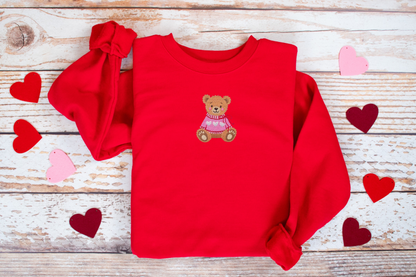 Red sweatshirt with a teddy bear design on a wooden surface with heart decorations.