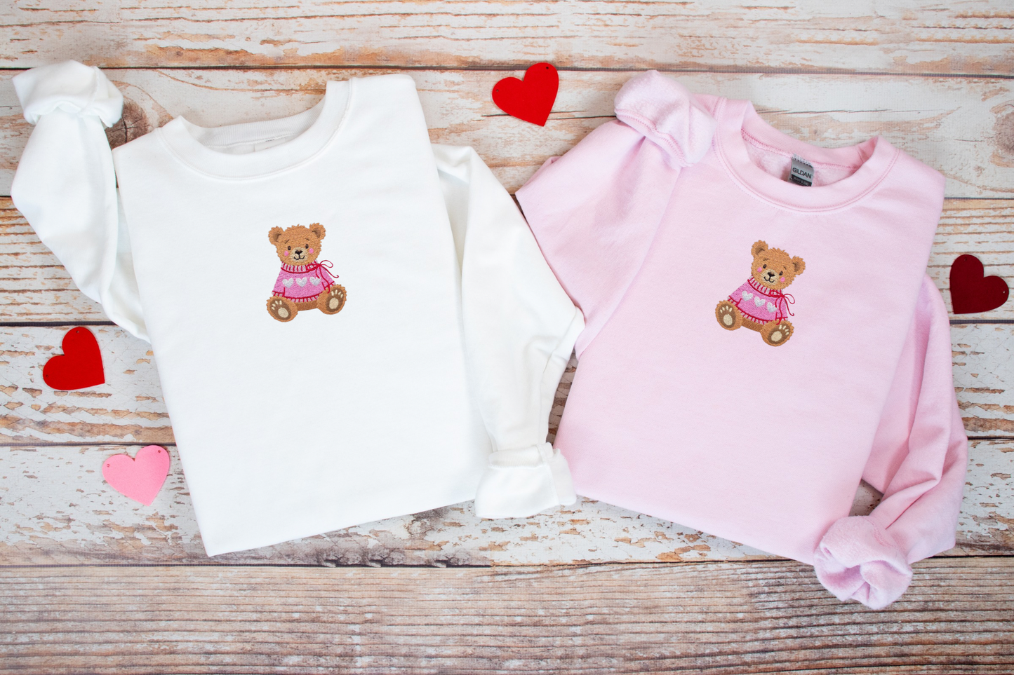 Two children's sweaters, one white and one pink, with teddy bear designs on a wooden surface.
