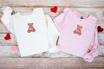 Two children's sweaters, one white and one pink, with teddy bear designs on a wooden surface.