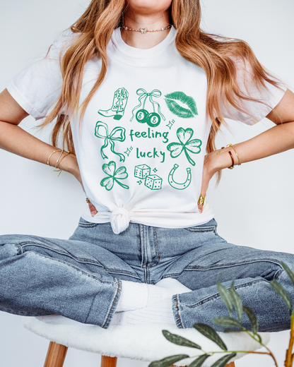 Person wearing a white t-shirt with green graphics and text, sitting on a chair. Tshirt says feeling lucky with line art design.
