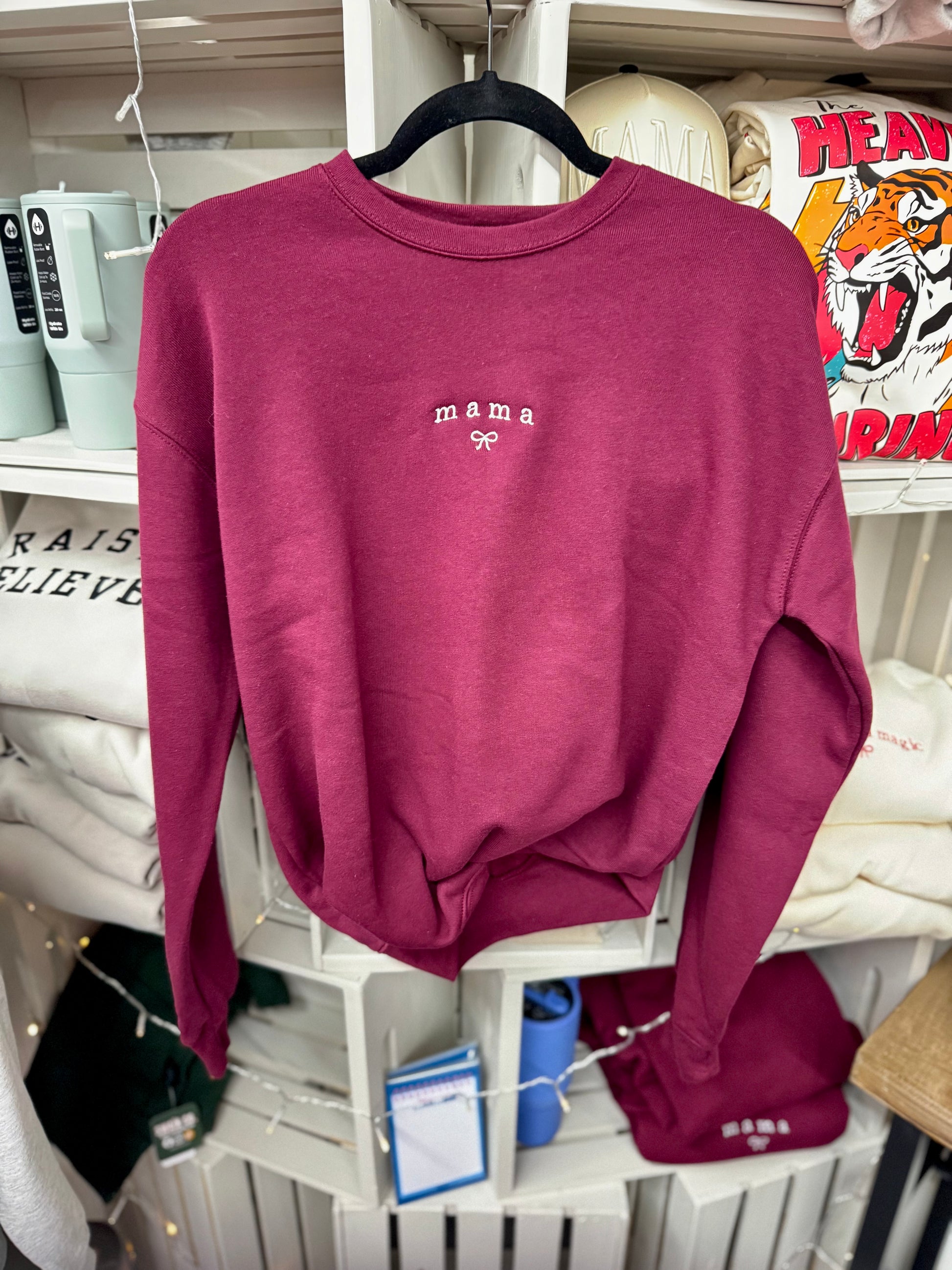 Maroon sweatshirt with 'mama' embroidery on a hanger against a store shelf background.