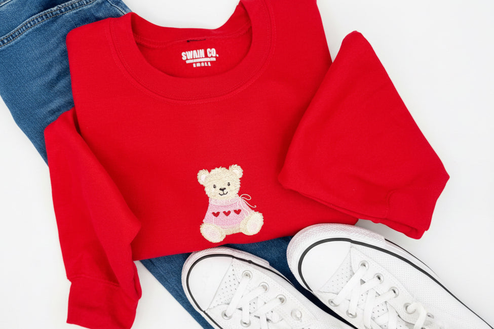 Red sweatshirt with teddy bear design, denim jeans, and white sneakers on a light gray background