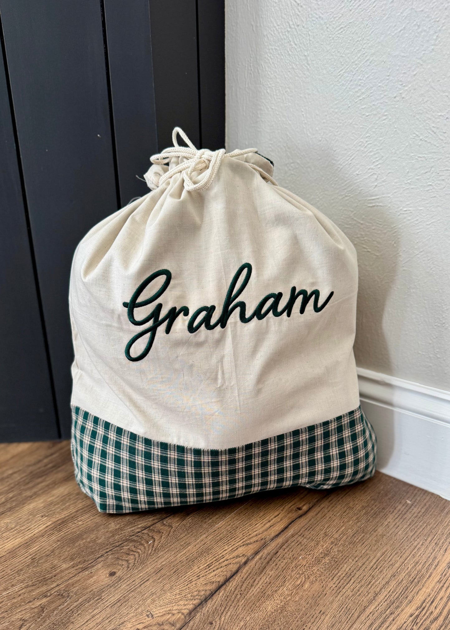 Drawstring bag with 'Graham' printed on it, sitting on a wooden floor against a white wall.