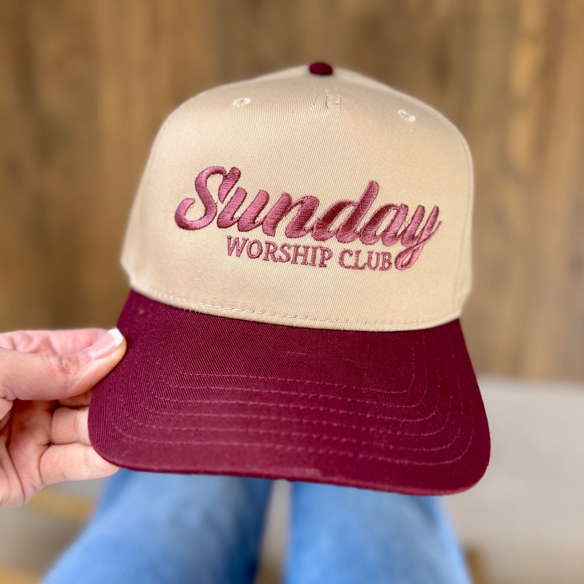 Beige and maroon cap with 'Sunday Worship Club' text held by a hand against a blurred background