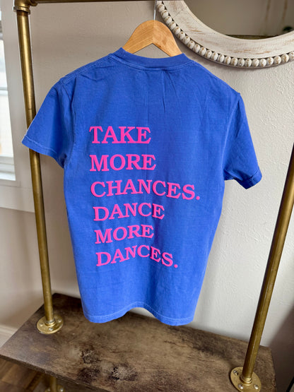 Blue t-shirt with pink text on a hanger against a neutral wall. Tshirt has puff print that says take more chances dance more dances.