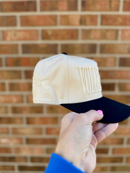 Beige and black cap held in front of a brick wall