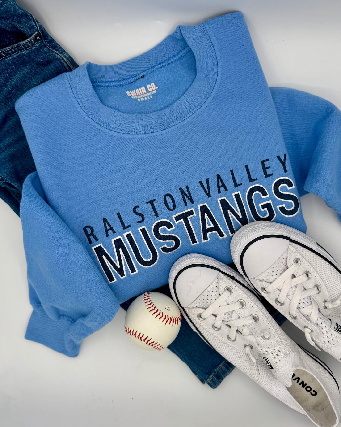 Blue sweatshirt with 'Ralston Valley Mustangs' text, white sneakers, and a baseball on a white surface.