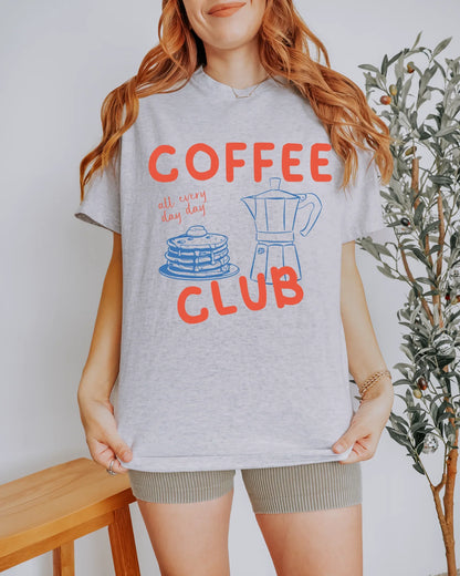 Person wearing a gray t-shirt with 'Coffee Club' design, standing indoors.