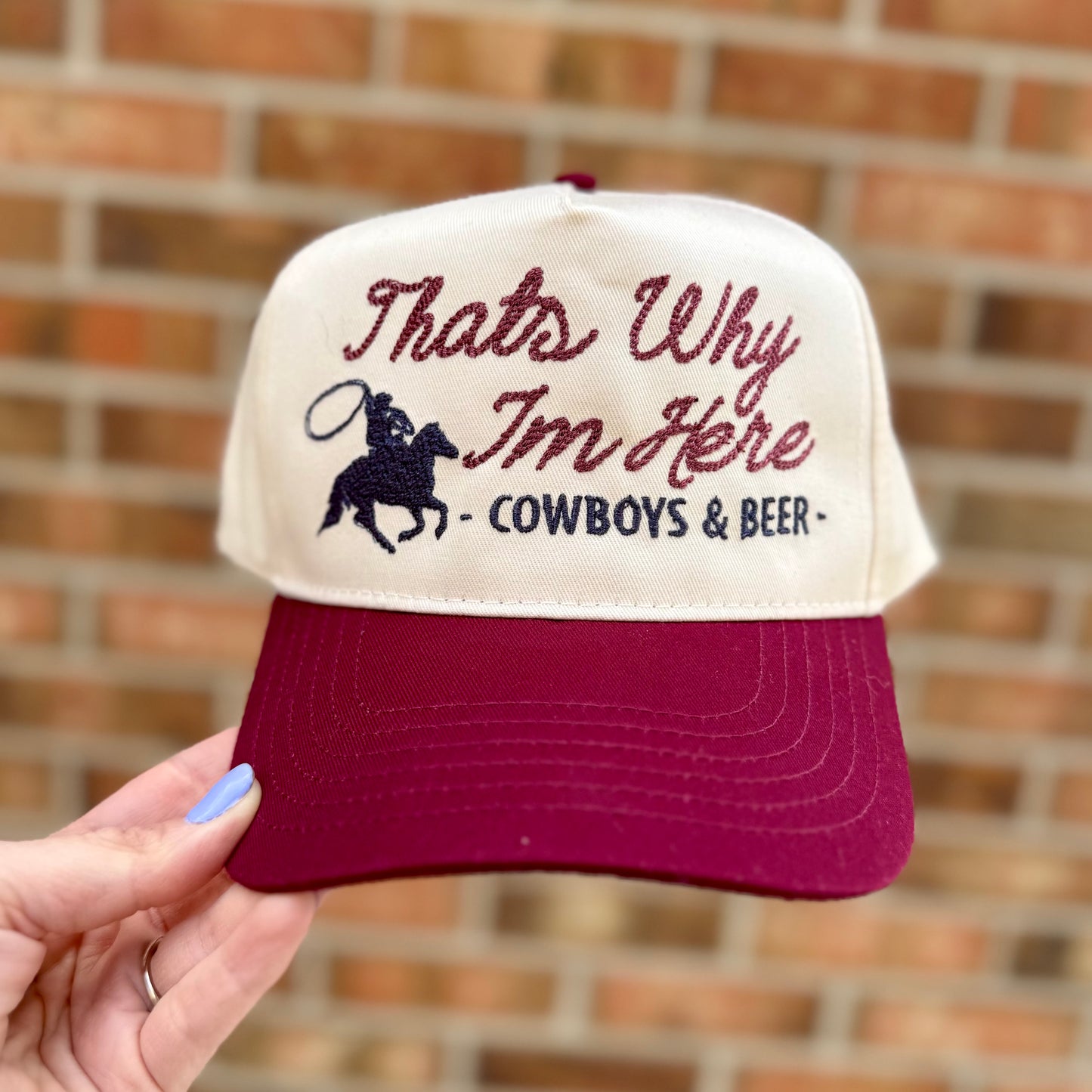 Cap with 'That's Why I'm Here - Cowboys & Beer' text held against a brick wall.