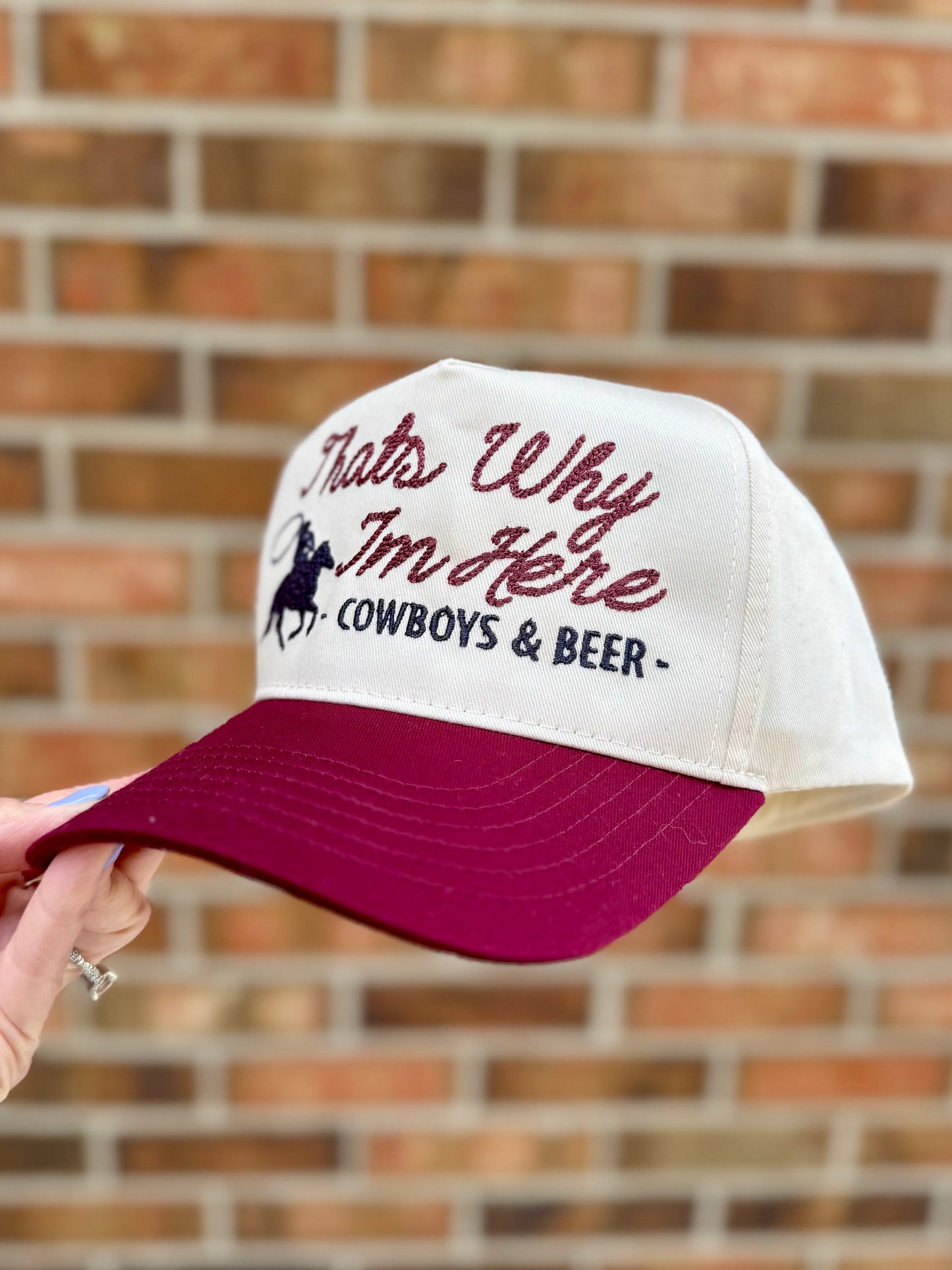Cap with 'That's Why I'm Here - Cowboys & Beer' text held against a brick wall.