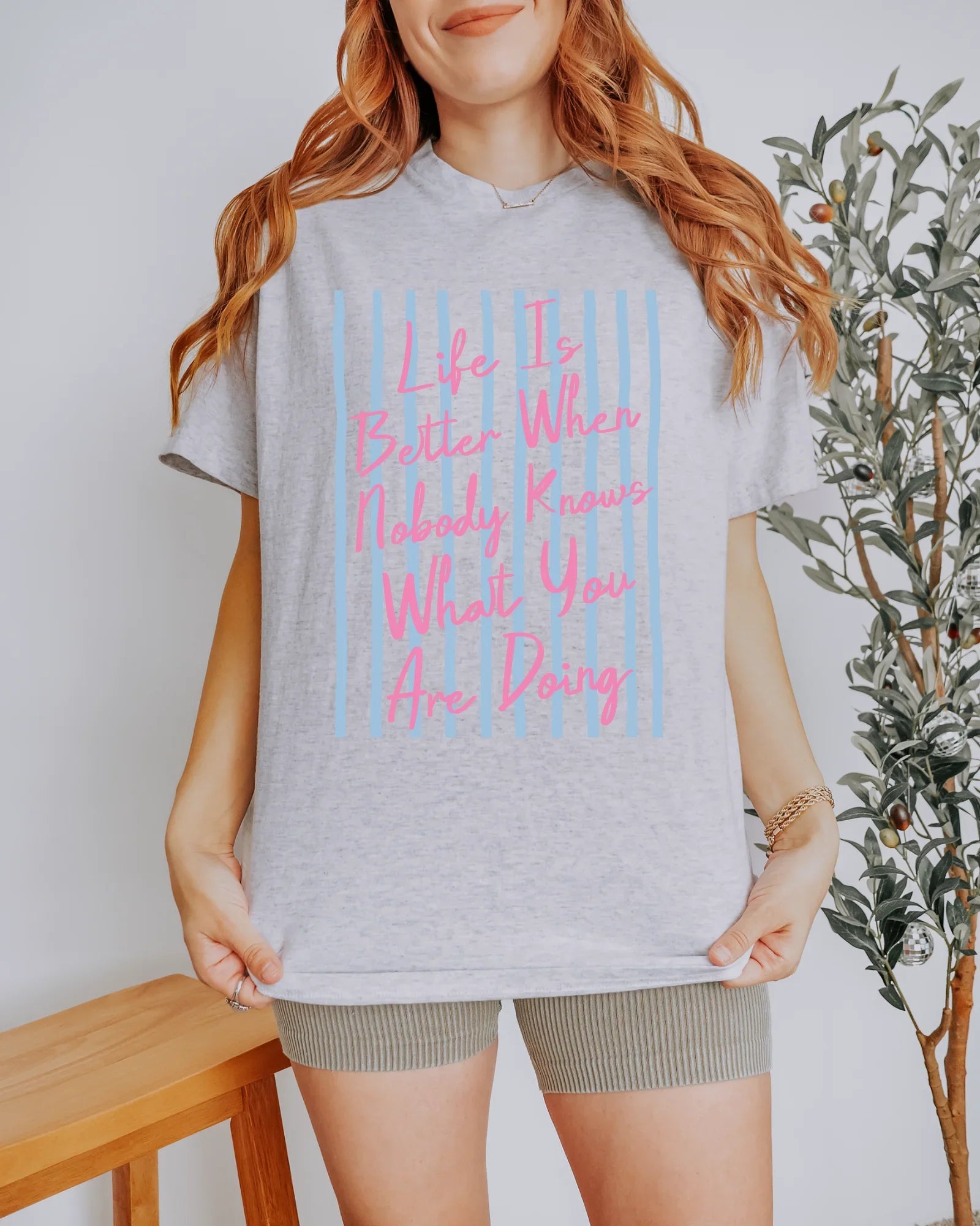 Person wearing a gray t-shirt with pink text, standing indoors next to a plant.